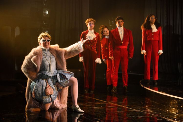 Actor playing Orlando kneeling in the front with a silk pastel blue tudor style costume with a large pink fluffy trench coat and white sunglasses. Ensemble in the background in red 1970's style suits with white shirts/ruffles